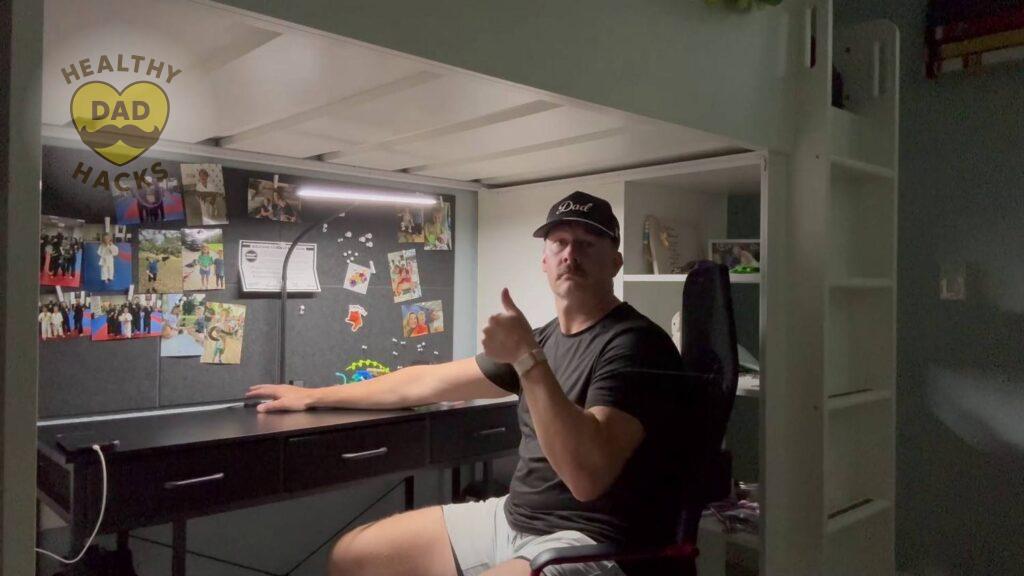 Dad sits at a desk under a loft bed, giving a thumbs-up next to the Best Gooseneck LED Desk Lamp with photos pinned behind him.