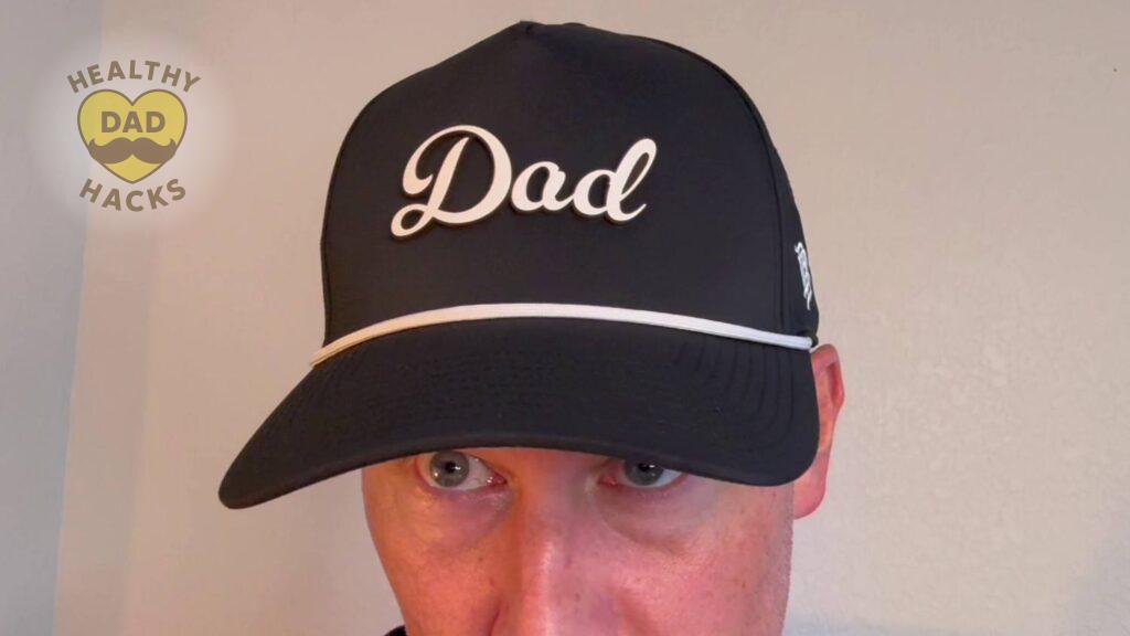 Review Dad wearing a black baseball cap with "Dad" in white cursive letters stands against a neutral-colored wall.