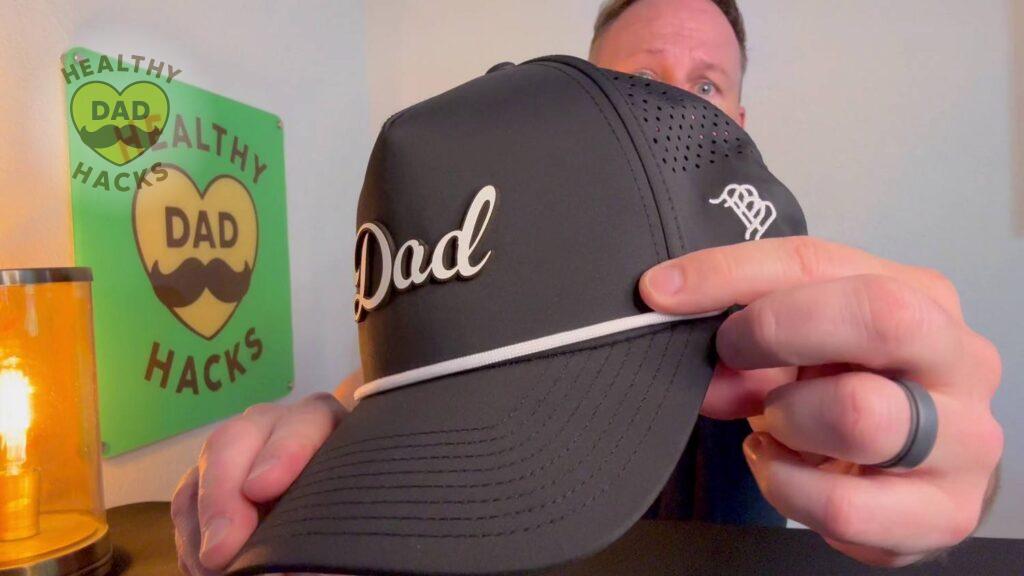 Dad points at a black cap with "Dad" written on it.